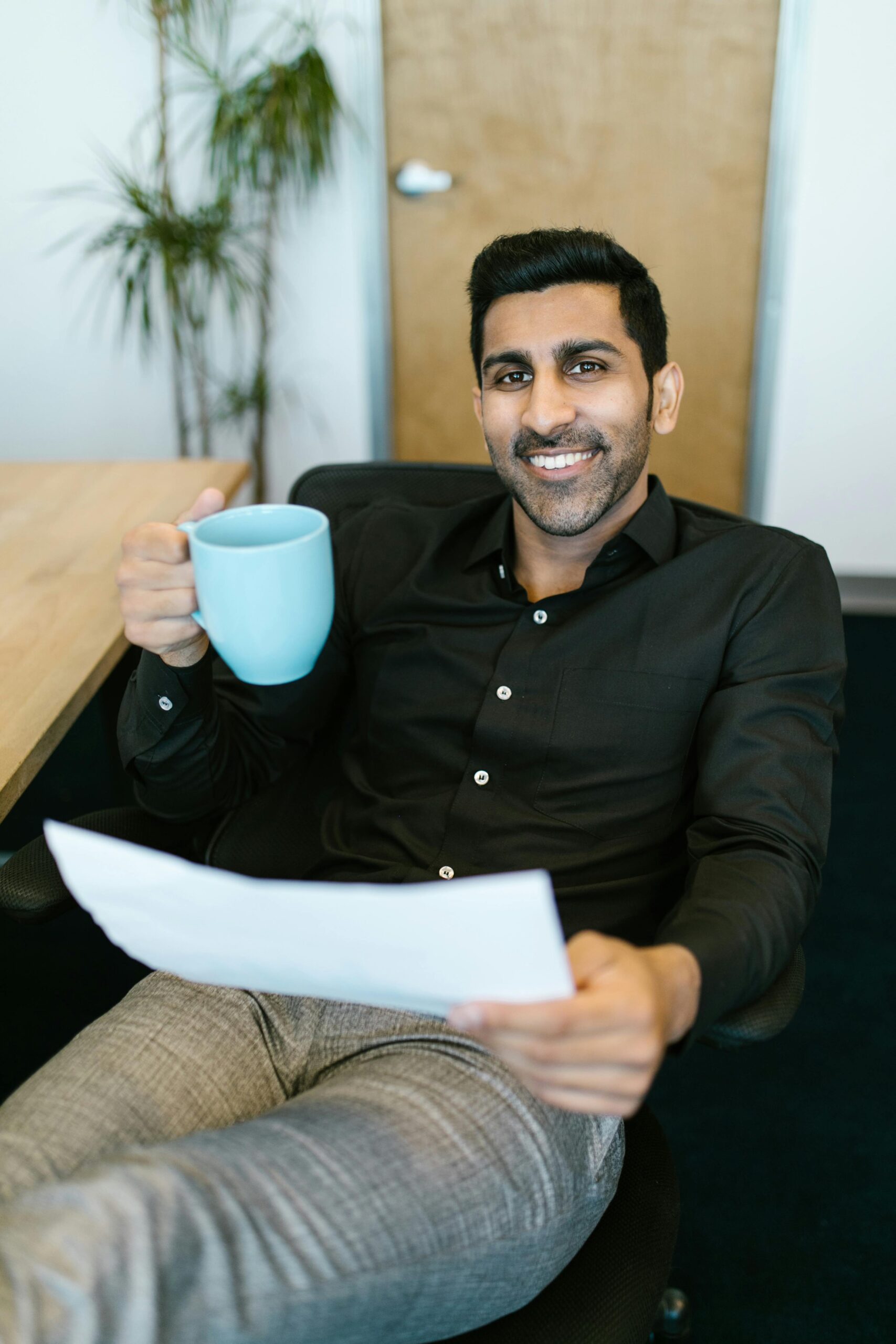 Businessman relaxing in office with a coffee, smiling confidently at the camera.